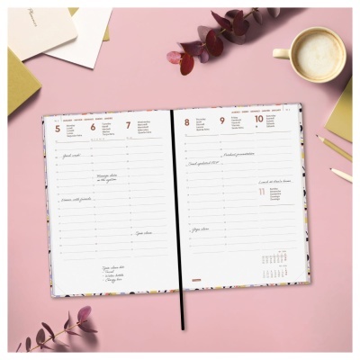 Agenda aberta com planejamento semanal, galhos, blocos de notas, lápis e chávena com bebida sobre fundo rosa.