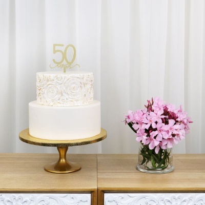 Bolo de aniversário branco de dois andares com topo dourado 50 anos e vaso de flores cor-de-rosa ao lado