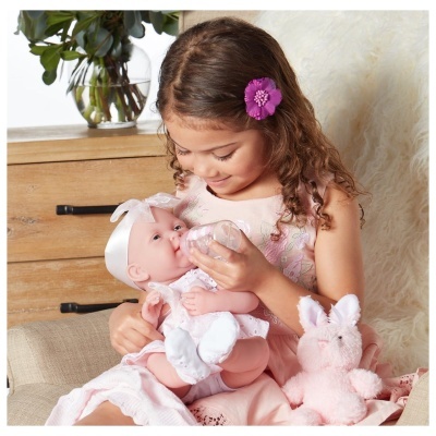 Criança a alimentar boneca de bebé com biberão, com peluche rosa ao lado.