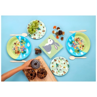 Conjunto de pratos descartáveis com desenhos de animais, talheres de madeira, guardanapo com coelho, uvas verdes e muffins com cobertura de chocolate numa mesa azul