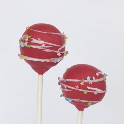 Dois cake pops vermelhos com confeitos coloridos e riscas de glacê branco em fundo branco
