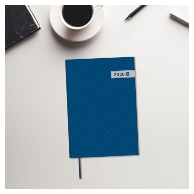 Agenda azul com etiqueta 2026 e marcador azul sobre mesa branca com café, caneta e livro.