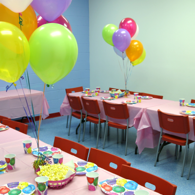 Mesas com toalhas cor-de-rosa, balões coloridos, pratos e copos coloridos, cadeiras vermelhas, parede azul e cinza