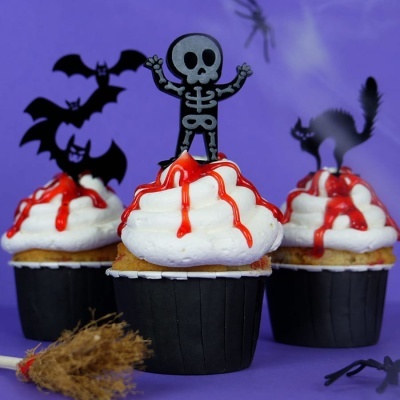 Cupcakes temáticos de Halloween com cobertura branca e calda vermelha, decorados com figuras de esqueleto, morcegos e gato preto