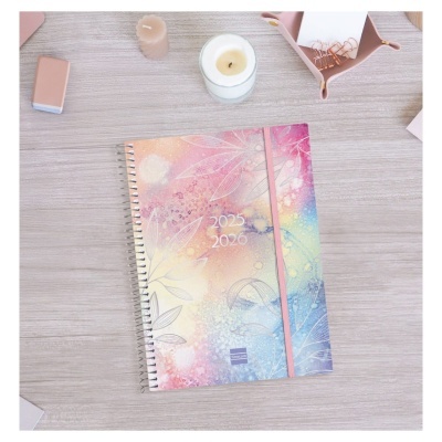 Agenda espiralada colorida 2025 2026 com padrão floral e elástico rosa numa mesa de escritório