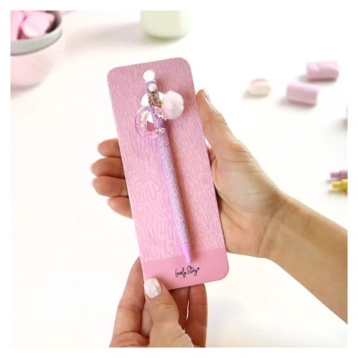 Caneta rosa decorada com anel brilhante e pompom, embalada em cartão rosa com texto.