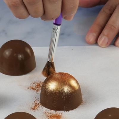 Bombons de chocolate semicirculares sendo decorados com pó dourado usando um pincel