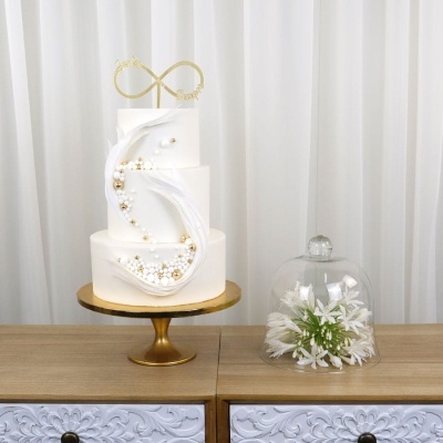 Bolo branco de três andares com decoração de pérolas e penas, suporte dourado e flores brancas em cúpula de vidro