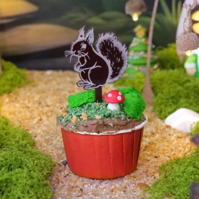 Cupcake decorado com glacê castanho, cogumelo vermelho de açúcar e topper de esquilo em cenário de floresta em miniatura