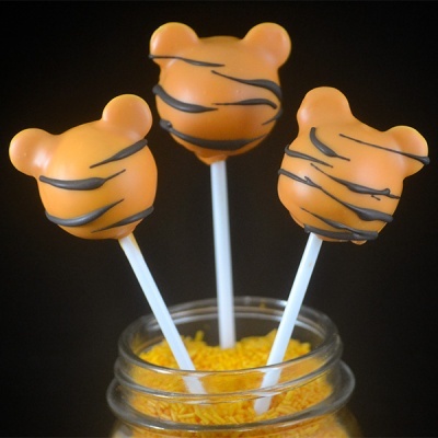 Três pirulitos de cabeça de urso laranja com riscas pretas em paus brancos dentro de frasco com granulados amarelos