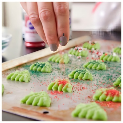 Mão polvilha açúcar vermelho sobre bolinhas verdes em forma de pinha numa bandeja de metal.
