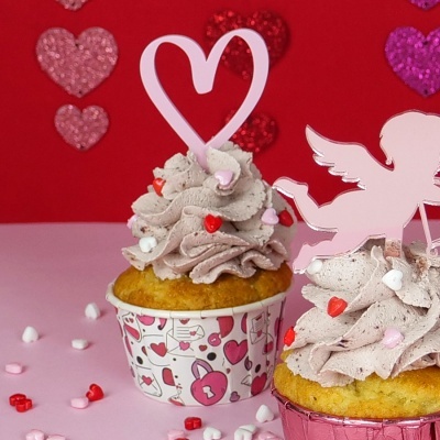 Cupcakes decorados com cobertura rosa e toppers de coração e anjo, em embalagens decoradas com corações, sobre mesa rosa e fundo vermelho com corações brilhantes.