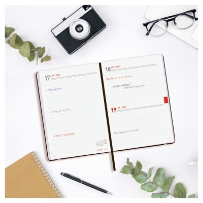 Agenda semanal aberta com caneta, caderno, óculos, câmera retro e folhas verdes sobre mesa branca