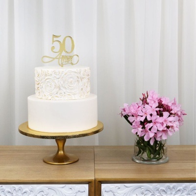 Bolo branco de dois andares com decoração de rosas e topo dourado 50 Anos, ao lado de vaso com flores cor-de-rosa sobre móvel de madeira