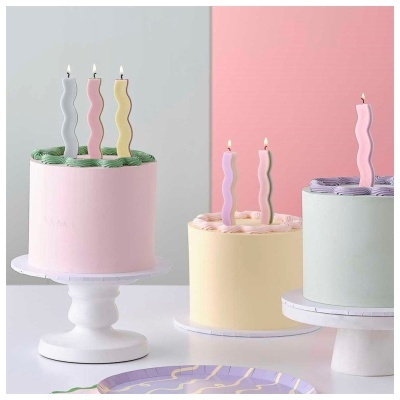Três bolos decorados com velas onduladas coloridas e acesas em fundo de cores suaves.