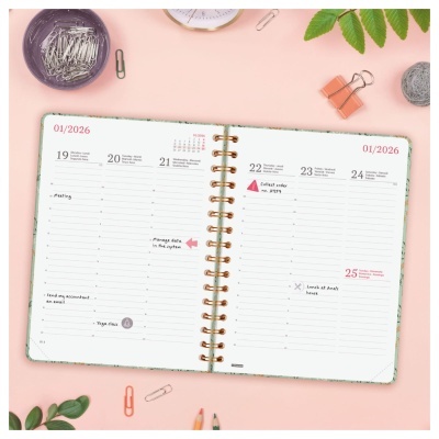 Agenda semanal aberta com espiral dourada sobre superfície rosa com clips e folhas verdes