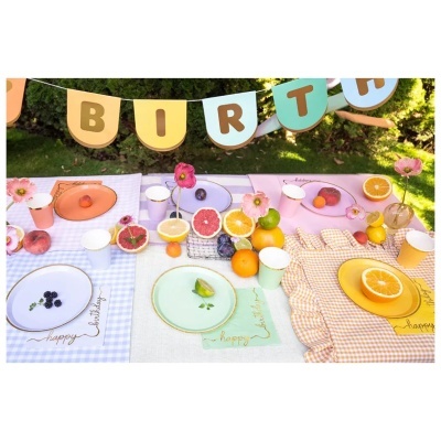 Bandeirolas com letras BIRTH e mesa de festa decorada com pratos coloridos, frutas e flores ao ar livre
