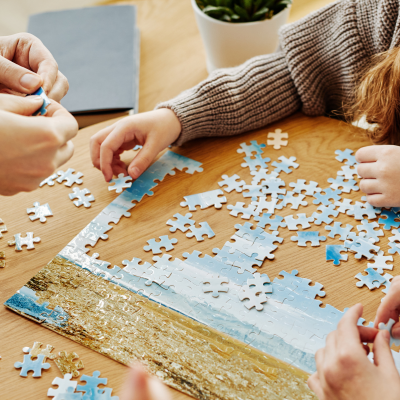 Três pessoas a montar um puzzle na mesa redonda de madeira com peças de céu azul e areia.