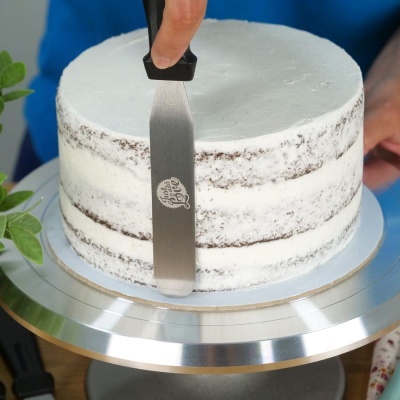 Bolo redondo branco com camadas castanhas numa base metálica e espátula com cabo preto a aplicar acabamento