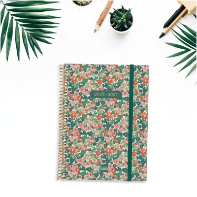 Agenda floral 2026-2027 com espiral dourada e elástico verde num fundo branco com folhas e canetas.