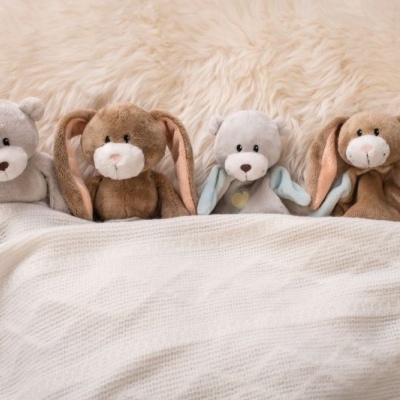 Quatro peluches, dois ursos cinza claro e dois coelhos castanhos, sob um cobertor creme numa cama com pele sintética