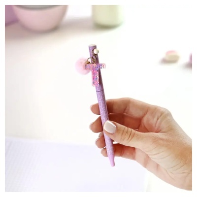 Caneta cor-de-rosa com pingente em forma de letra T e pompom, segurada por uma mão.