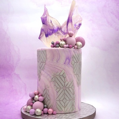 Bolo cilíndrico lilás com decoração geométrica prateada e bolas coloridas no topo