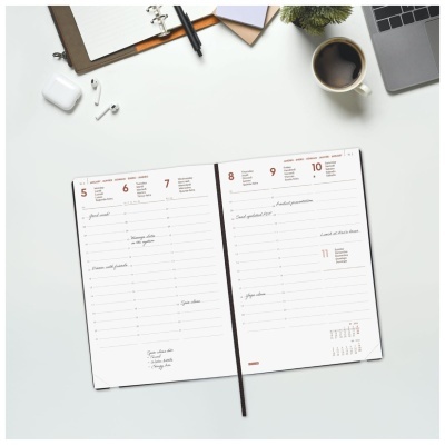 Agenda semanal aberta, caneca de café, portátil, caderno, caneta, auriculares sobre mesa branca.