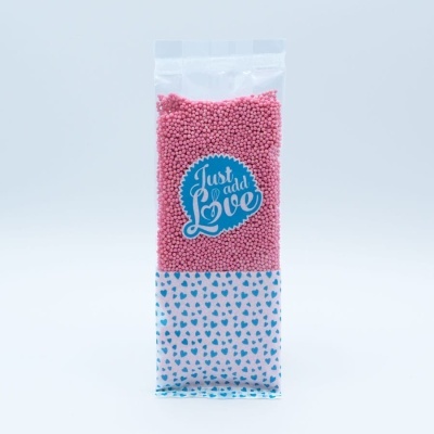 Pacote transparente com bolinhas cor-de-rosa e etiqueta 'Just add Love', fundo branco com corações azuis.