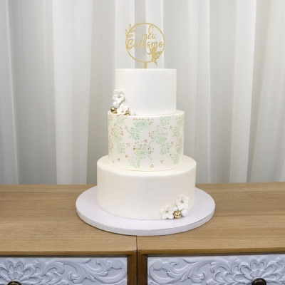 Bolo branco de três andares decorado com flores, bolinhas douradas e padrão verde, com topo dourado escrito O meu Batismo