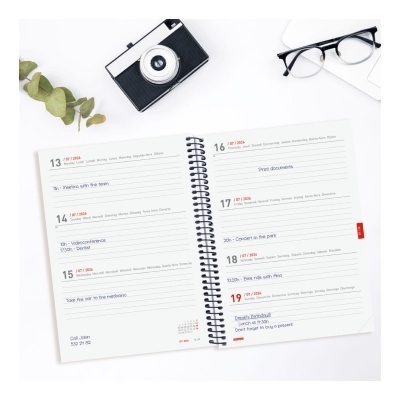 Agenda semanal aberta com espiral preta, câmara fotográfica retro, óculos e folhas de eucalipto sobre mesa branca