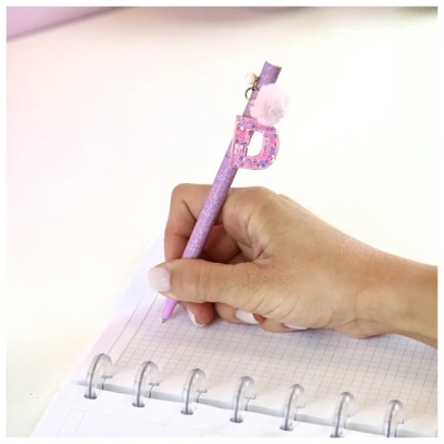 Caneta cor-de-rosa com pendente letra D e pompom numa mão escrevendo num caderno quadriculado com argolas