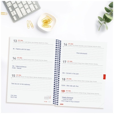 Agenda semanal aberta com clips dourados, teclado cinzento e ramo verde sobre fundo branco