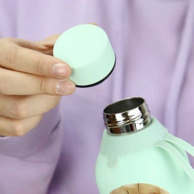 Garrafa térmica verde água com tampa removível e interior metálico, segurada por pessoa com capuz lilás