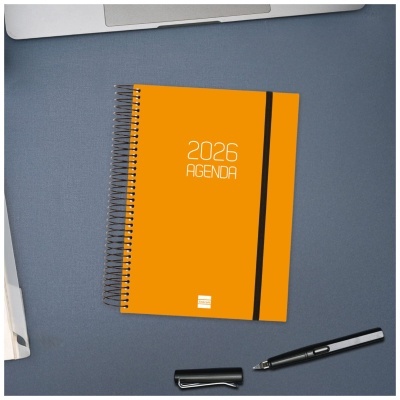 Agenda 2026 laranja com espiral e elástico preto sobre mesa cinzenta ao lado de portátil e caneta preta