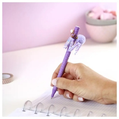 Caneta roxa com adorno letra M, pompom e acessórios dourados usada para escrever em caderno quadriculado