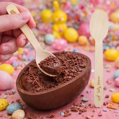 Ovo de chocolate meio amargo com granulado, colher de madeira gravada e ovos de páscoa coloridos em fundo rosa