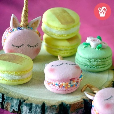 Macarons coloridos decorados com temas de unicórnio e flores sobre base de madeira com fundo rosa