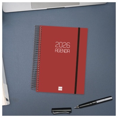 Agenda vermelha 2026 com espiral e elástico preto sobre superfície azul com caneta e parte de laptop