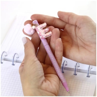 Caneta rosa decorada com letra E, pompom e contas nas mãos sobre caderno quadriculado