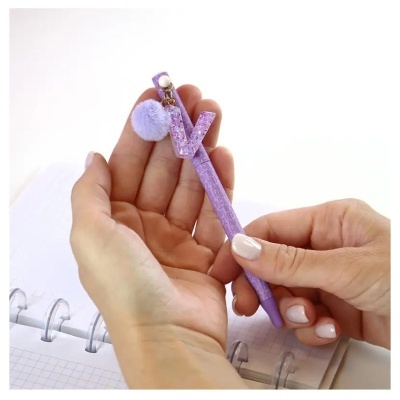 Caneta roxa decorada com letra V e pompom lilás segura por mãos sobre caderno