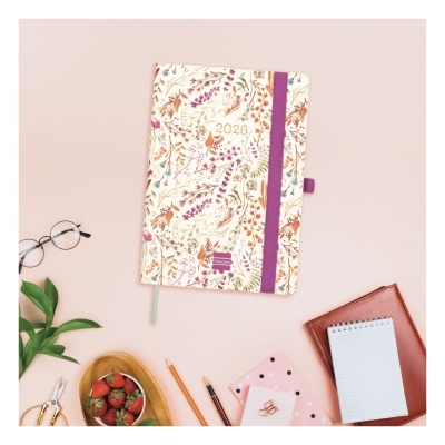 Agenda 2026 floral com elástico roxo e marcador verde num ambiente com óculos, planta, morangos, canetas, estojo cor-de-rosa e bloco de notas.