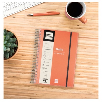 Caderno planner diário laranja com caneta, teclado, planta e caneca de café numa mesa de madeira.