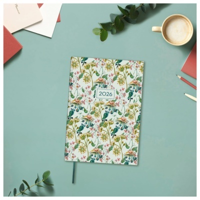 Agenda 2026 com padrão floral colorido sobre mesa azul clara com canetas, ramos verdes e chávena de café.