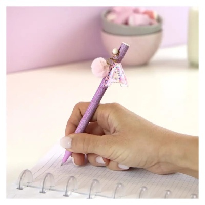 Caneta rosa decorada com letra A e pompom, segurada por mão escrevendo em caderno quadriculado.