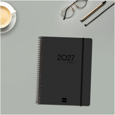Agenda preta espiralada com o ano 2027 e acessórios de escrita junto a uma chávena de café.