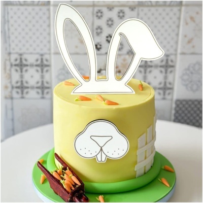 Bolo amarelo decorado com temática de coelho, incluindo orelhas e focinho de papel, mini cenouras e base verde.