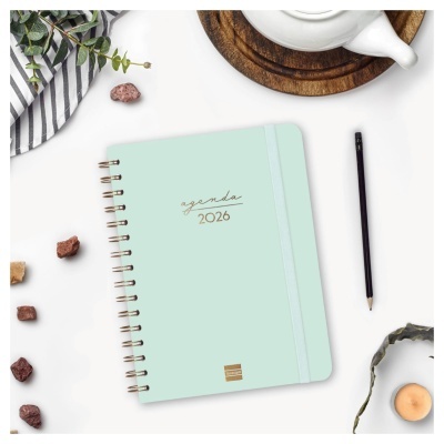 Agenda verde claro 2026 com elástico e argolas douradas numa mesa branca com objetos decorativos