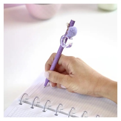 Caneta roxa com adereço letra C e pompom, sendo usada para escrever em caderno quadriculado.