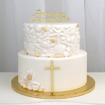 Bolo de comunhão branco com conchas em relevo, texto dourado Primeira Comunhão e cruz dourada